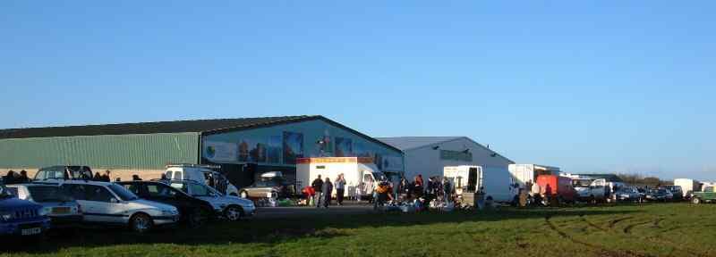 Cars, seller stalls and shoppers outside the large market shed at Môna Car Boot Sale. Attribution: Anglesey Info. Source: https://www.anglesey.info/mona-car-boot/