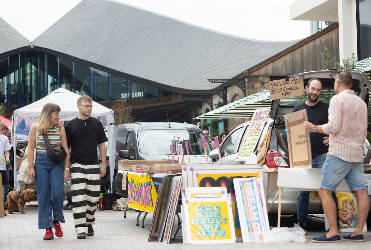King's Cross official event photograph showing artists selling colourful prints from open car boots as shoppers browse Art Car Boot Fair at Lewis Cubitt Square. Source: https://www.kingscross.co.uk/media/P_KXC_ECA_CBS_N2625_kxweb-1240x836.jpg?cache=1755009076