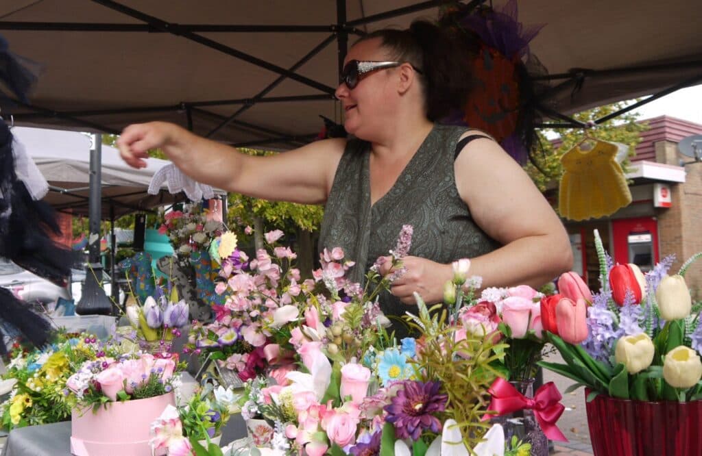 A trader serving visitors from a flower and craft stall at Staveley Market Source: https://staveleytowncouncil.gov.uk/wp-content/uploads/2024/03/xmarket-4-charlotte-scaled-e1710511517838-1024x666.jpg.pagespeed.ic.oGzJ-Hdv_v.webp