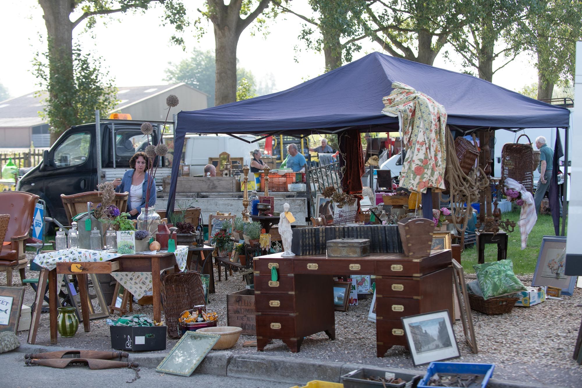 Outdoor antiques stall with shoppers and seller vehicles at Shepton Flea Source: https://www.sheptonflea.com/wp-content/uploads/2023/05/850_2641-2.jpg